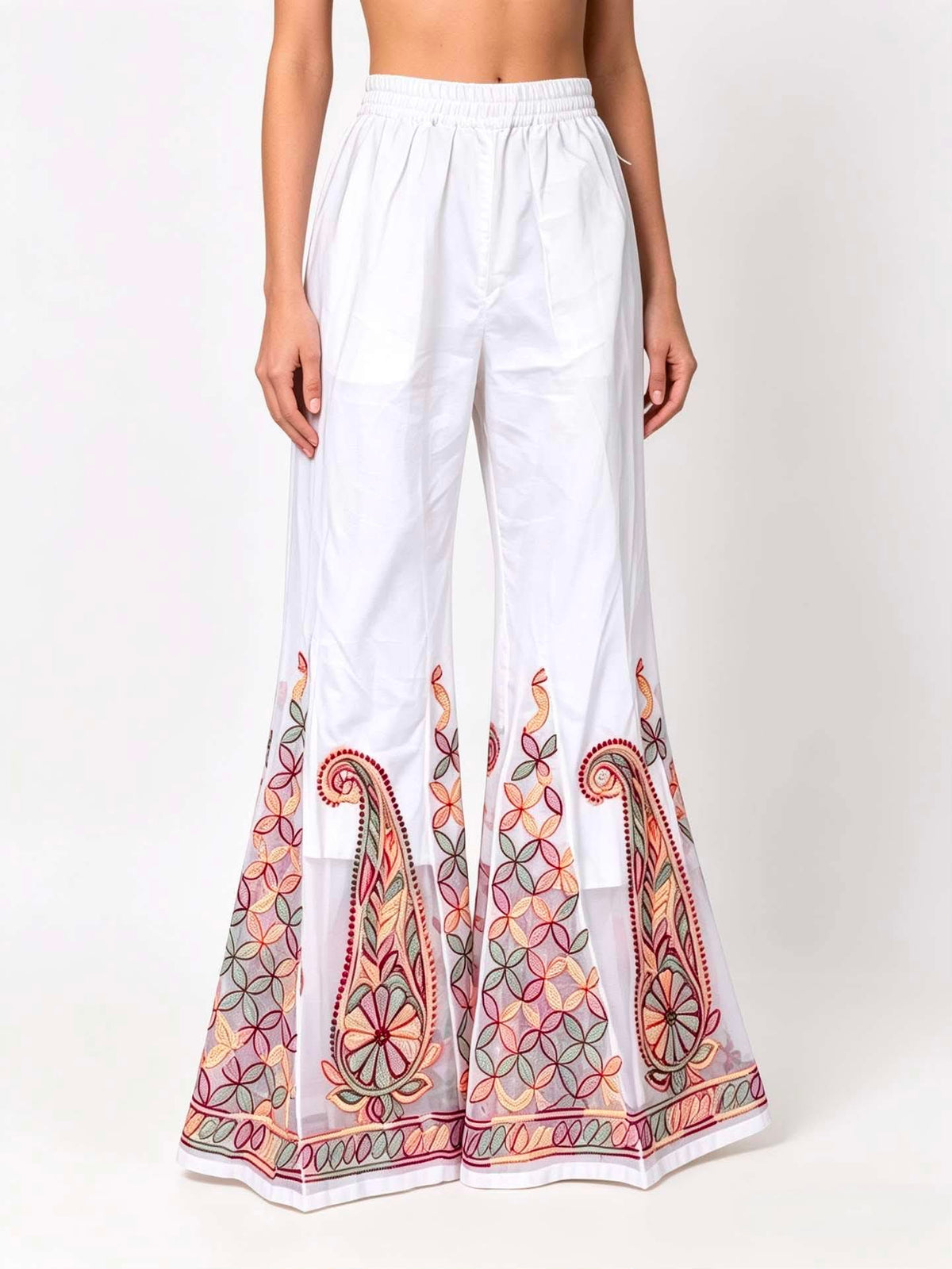 A front view of a model wearing high-waisted white semi-georgette sharara pants with a fluid, wide-leg flared silhouette, featuring a sheer white organza panel at the bottom hem decorated with intricate multi-color traditional paisley and geometric embroidery in rust red, sage green, and pastel yellow.