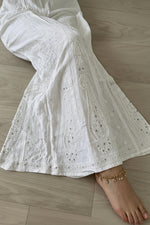 White embroidered cotton flare pants with eyelet detailing, shown on a model lying on a light wood floor.