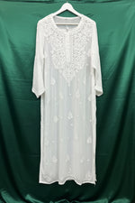 White embroidered long kurti dress with floral lace detailing on the chest and scattered embroidered motifs on the skirt, hanging on a white hanger against a black background.