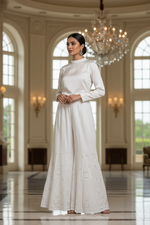A woman stands in a grand, elegant hall wearing a long-sleeved white maxi dress with a high mock neck and subtle eyelet embroidery near the hem. The background features arched windows, warm wall sconces, a large crystal chandelier, and a polished floor.