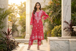 Woman in a red embroidered outfit standing in an outdoor setting with plants and architecture.
