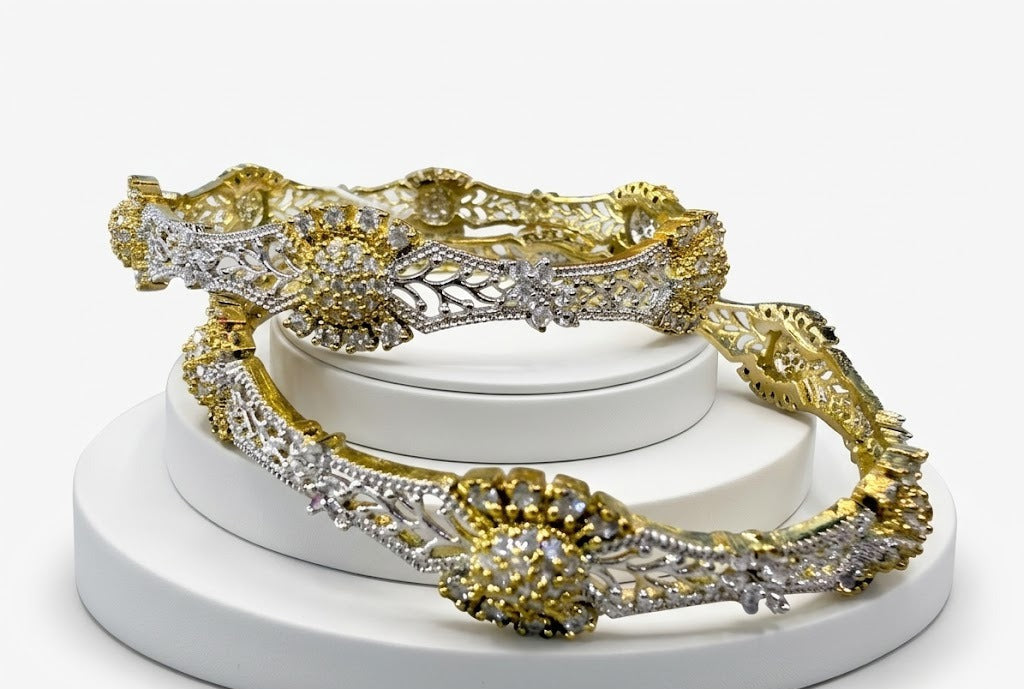 Pair of Zahra dual-tone bangles in gold and silver plating, featuring an intricate wavy floral lattice design and large starburst American Diamond clusters on a white stand.