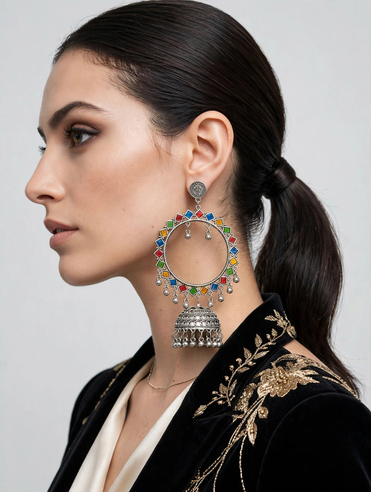 A woman in profile wearing a large silver-toned jhumka-style earring adorned with colorful red, blue, and green enamel paint, paired with a black embroidered jacket.