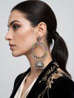 A woman in profile wearing a large silver-toned jhumka-style earring adorned with colorful red, blue, and green enamel paint, paired with a black embroidered jacket.