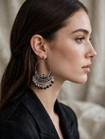 A close-up side profile of a woman wearing a large silver-toned huggie hoop earring adorned with dangling black beads and metallic fringe, set against a soft beige background.