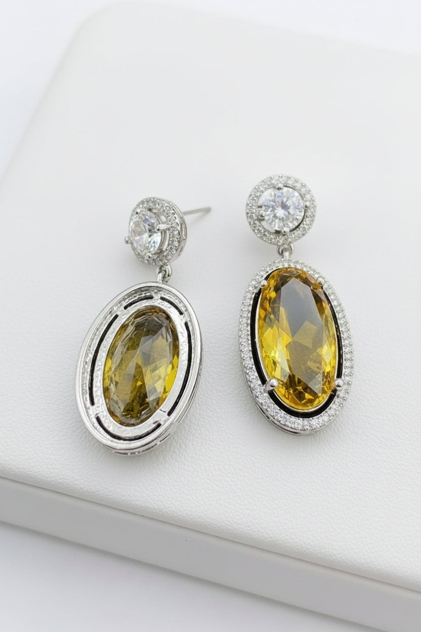 A pair of silver-tone drop earrings with large oval yellow gemstones, displayed on a white surface next to a gray card and small clear stud earrings.