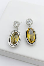 A pair of silver-tone drop earrings with large oval yellow gemstones, displayed on a white surface next to a gray card and small clear stud earrings.