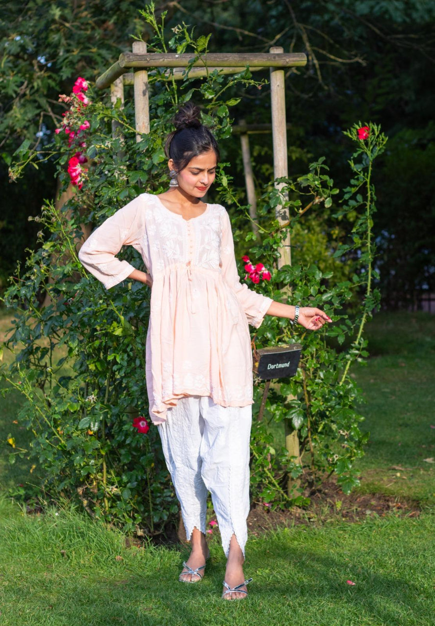A woman standing outdoors wearing a short frock style kurti with embroidery and a tie-up waist, in a garden showing full look with trousers