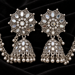 A pair of ornate silver-toned Indian jhumka earrings with intricate filigree work, clear stone inlays, and dangling bead chains, displayed on a light beige background.