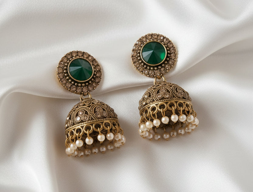 A pair of ornate gold-toned jhumka earrings with green oval stones, displayed on a gold stand with measurement labels showing drop length, jhumka width, and weight.