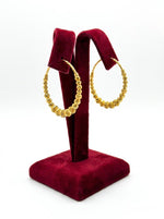 A pair of large gold hoop earrings with textured, beaded detailing, displayed on a dark red velvet jewelry stand against a white background.