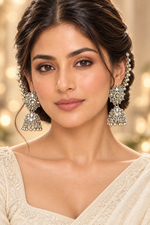 A close-up portrait of a woman wearing ornate silver-toned jhumka earrings with intricate crystal embellishments and a matching hair ornament, paired with a cream-colored embroidered traditional garment against a warm beige background.