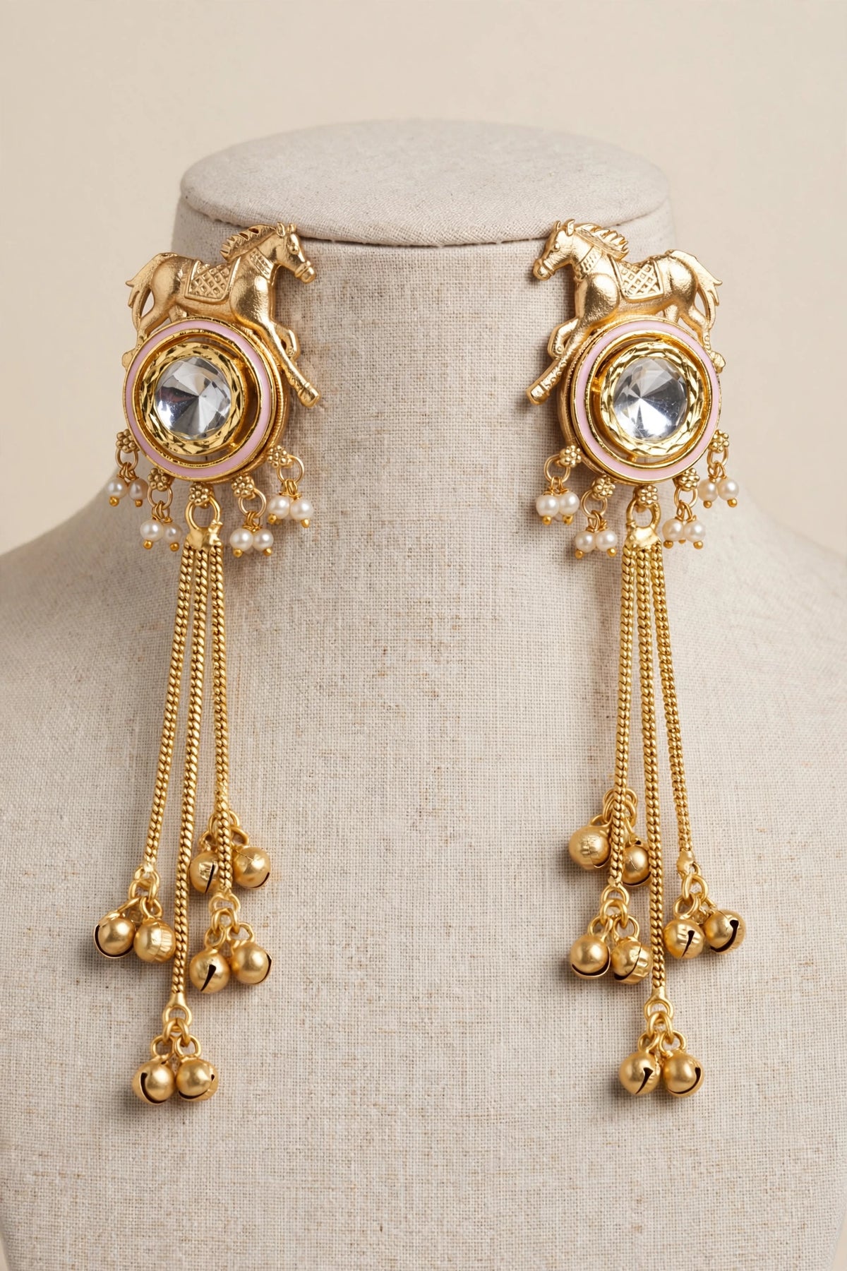 Rose Pink Golden Gallop horse motif earrings with reflective kundan centres, pearl-style beads and gold-tone tassels on a linen bust.