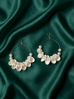 Ivory white shell-shaped hoop earrings with faux pearls and fishhook ear wires on green fabric.