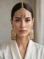 A woman with her hair pulled back wears an ornate gold-toned Indian-style maang tikka with colorful dangling beads and matching long earrings, paired with a white satin robe against a soft beige background.