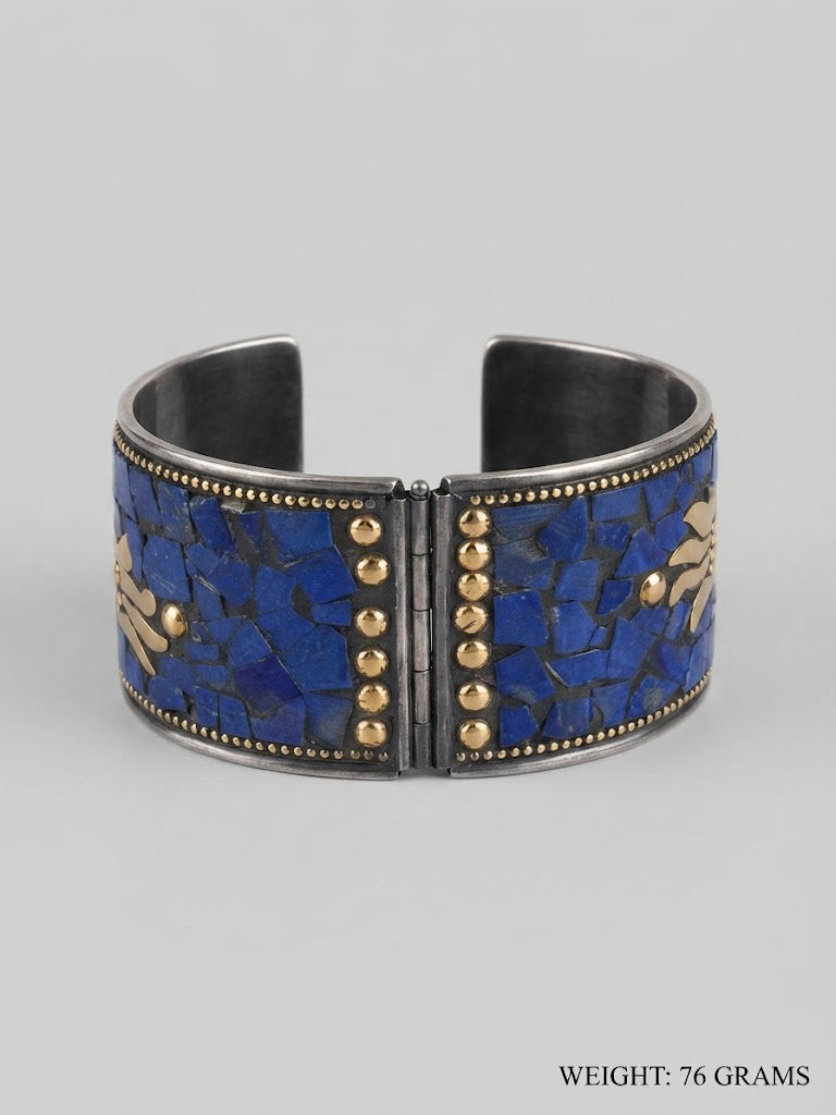 A wide silver-toned cuff bracelet featuring a mosaic inlay of deep blue lapis lazuli-style stones and small gold-tone studs, shown on a light gray background. Text in the lower right corner reads 'WEIGHT: 76 GRAMS'.