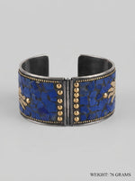 A wide silver-toned cuff bracelet featuring a mosaic inlay of deep blue lapis lazuli-style stones and small gold-tone studs, shown on a light gray background. Text in the lower right corner reads 'WEIGHT: 76 GRAMS'.