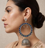 A close-up side profile of a woman wearing a large, ornate silver-toned dangle earring embellished with deep blue stones and silver bell-shaped drops, paired with a beige embroidered garment.