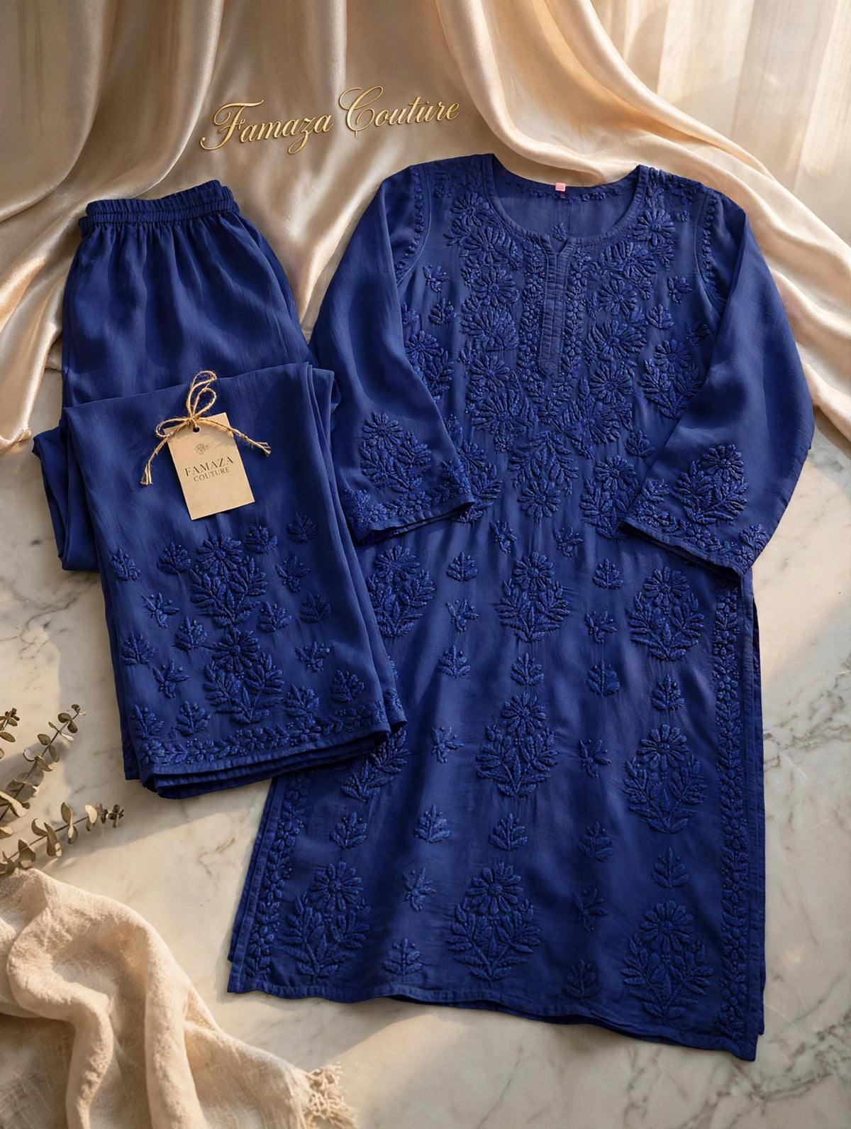 A royal blue embroidered tunic and matching pants set laid flat on a marble surface with draped cream satin fabric. The tunic has long sleeves, a round neckline, and detailed floral embroidery across the front.