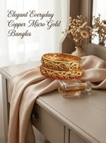 A set of two micro gold-plated copper bangles with an intricate, open-work floral and vine pattern, elegantly displayed on a champagne-colored silk cloth next to a glass perfume bottle on a light wooden vanity.