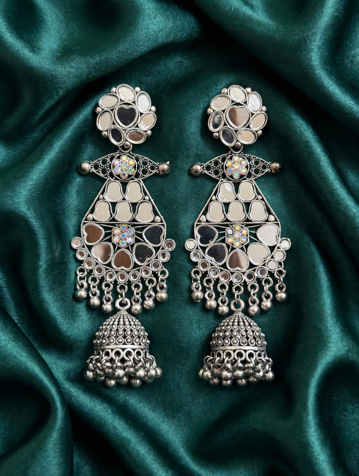 Mirror Muse silver-tone mirror jhumka earrings laid on green satin, with floral tops, iridescent crystal accents and ornate bell drops