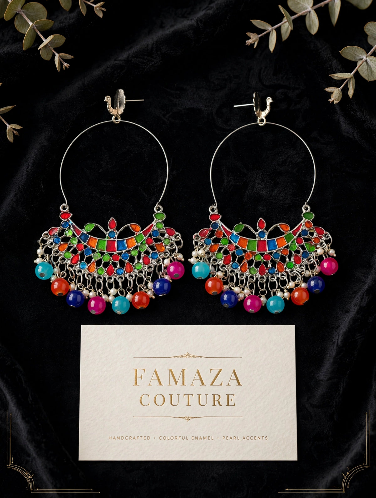 A pair of large silver-tone hoop earrings with colorful enamel and bead detailing, displayed on a black velvet background with decorative leaves and a branded card reading FAMAZA COUTURE.