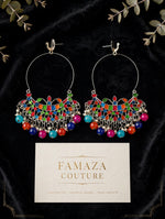 A pair of large silver-tone hoop earrings with colorful enamel and bead detailing, displayed on a black velvet background with decorative leaves and a branded card reading FAMAZA COUTURE.
