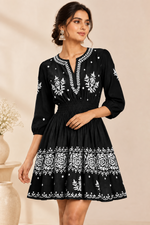 A woman modeling a black short-sleeve mini dress with white floral embroidery, featuring a split neckline, smocked waist, and a flared skirt, set against a warm beige background.