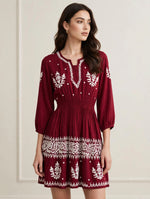 A woman wearing a maroon mini dress with white embroidered patterns, standing against a light beige wall.