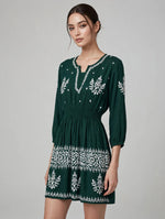 A woman modeling a bottle green mini dress with white embroidered patterns, featuring a split neckline, 3/4 sleeves, and a gathered waist, set against a plain light gray studio background.