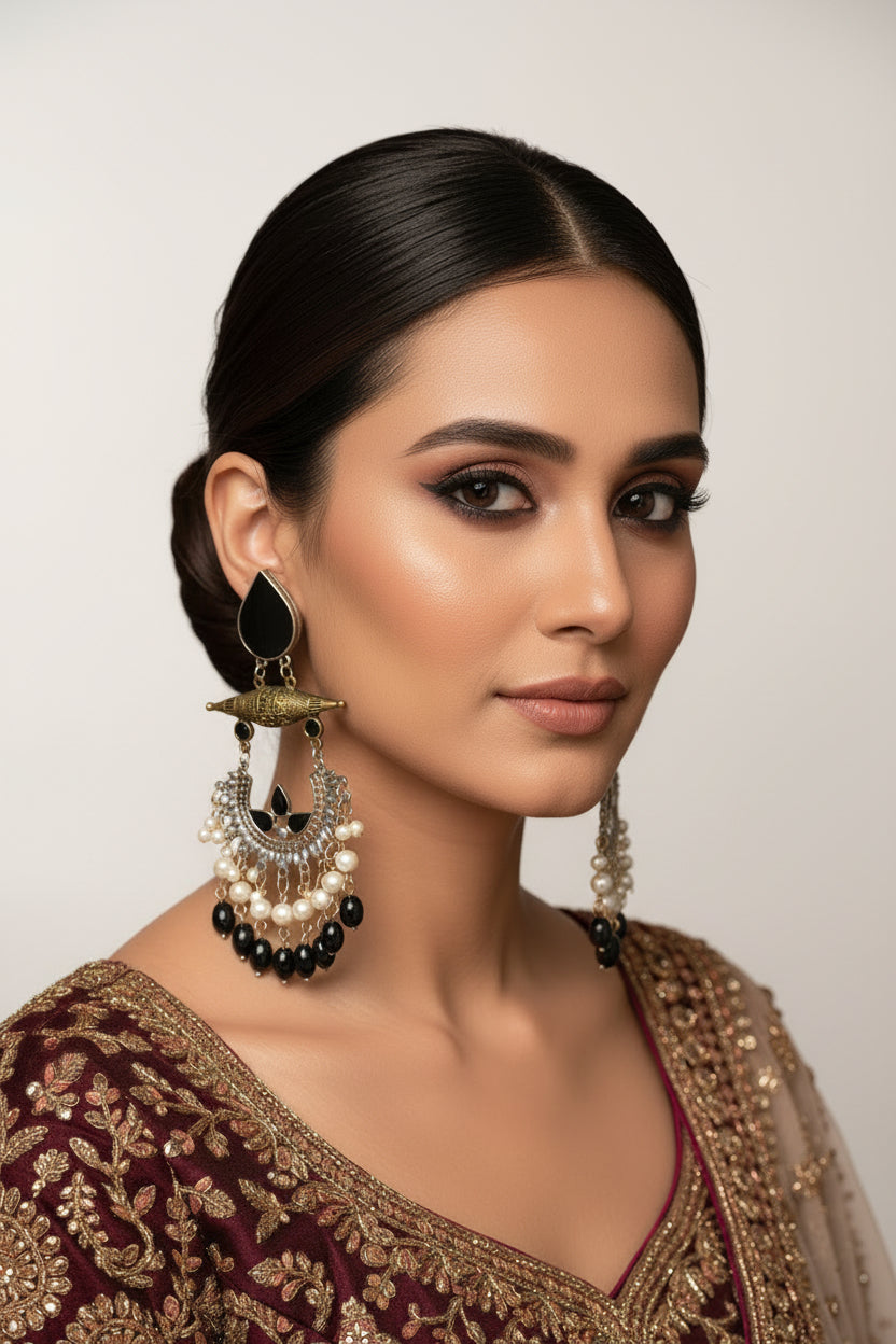 A pair of ornate Indian jhumka earrings displayed on a white faux fur background with measurement and weight labels. The earrings feature a black teardrop stone, an antique gold-toned top piece, a silver-toned crescent-shaped lower piece with black stones, and dangling white and black pearl chains.