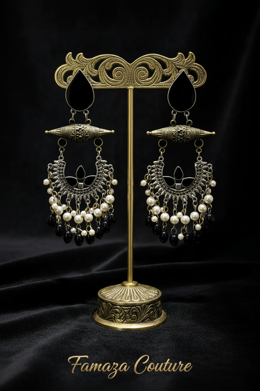 A pair of ornate traditional-style dangle earrings displayed on a black velvet surface. The earrings feature a gold-toned filigree cone bar, silver-toned crescent-shaped metalwork, white pearl beads, and dangling black faceted beads.