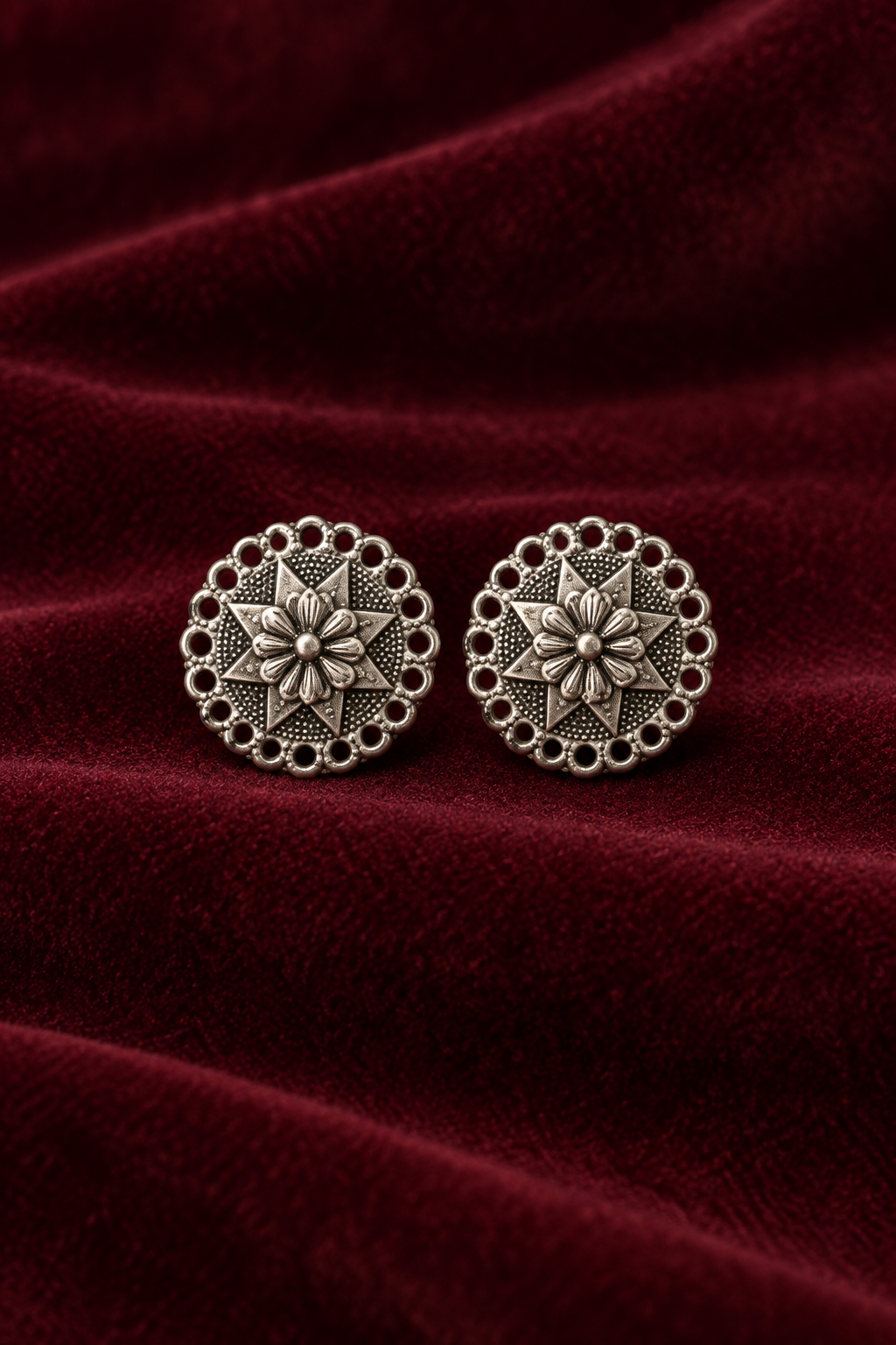 A pair of round silver-toned earrings with an intricate floral starburst design and scalloped border, displayed on a draped deep burgundy velvet fabric.
