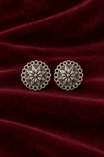 A pair of round silver-toned earrings with an intricate floral starburst design and scalloped border, displayed on a draped deep burgundy velvet fabric.