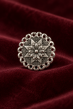 A round silver-toned metal button with an intricate floral starburst pattern and scalloped border, displayed on a draped dark red velvet fabric background.