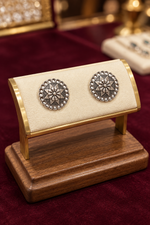 A pair of round silver-toned earrings with intricate floral starburst motifs and small clear stones, displayed on a cream and gold stand atop a wooden base against a dark red velvet background.