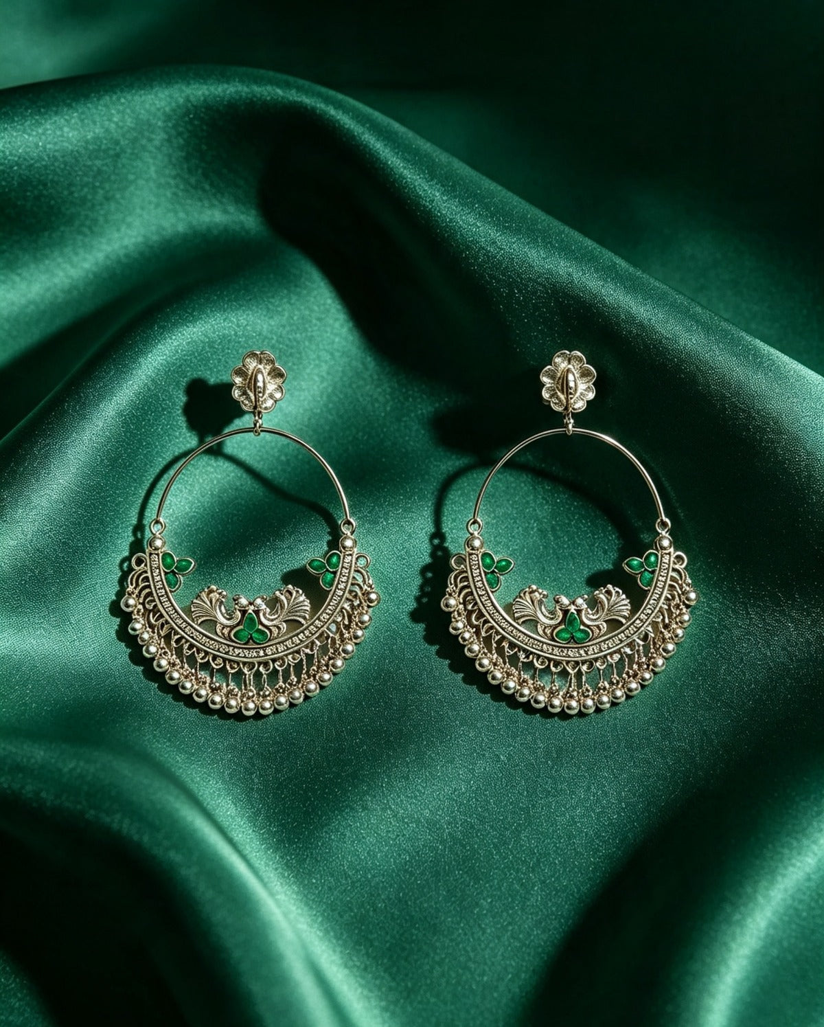 Pair of oxidized silver peacock chandbali hoop earrings with emerald green stone accents and ghungroo fringe — displayed on rich emerald green satin silk, antique silver finish, luxury ethnic jewelry by Famaza Couture.