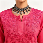 A close-up of a person wearing a bright pink embroidered kurta with floral embroidery and a silver-toned ornate choker necklace.