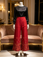 A mannequin wearing a black satin long-sleeve top and high-waisted red wide-leg trousers with gold floral embroidery near the hems, displayed in front of a brown velvet sofa with gold trim in an elegant interior.