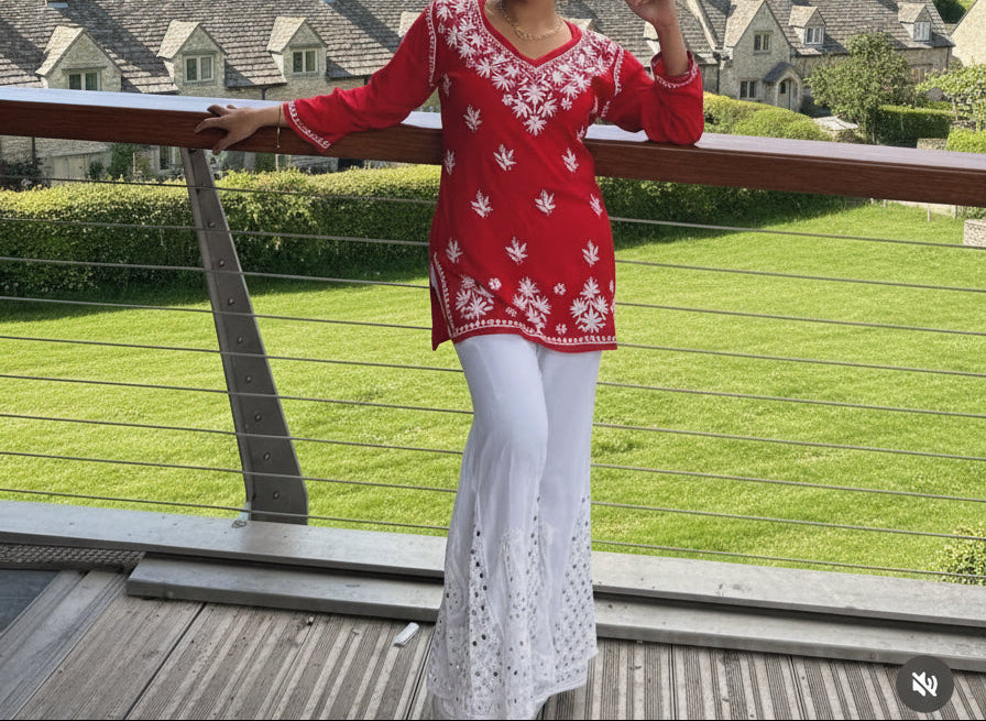 Woman in a red top with white patterns and white pants standing by a railing with a cityscape background.