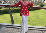 Woman in a red top with white patterns and white pants standing by a railing with a cityscape background.