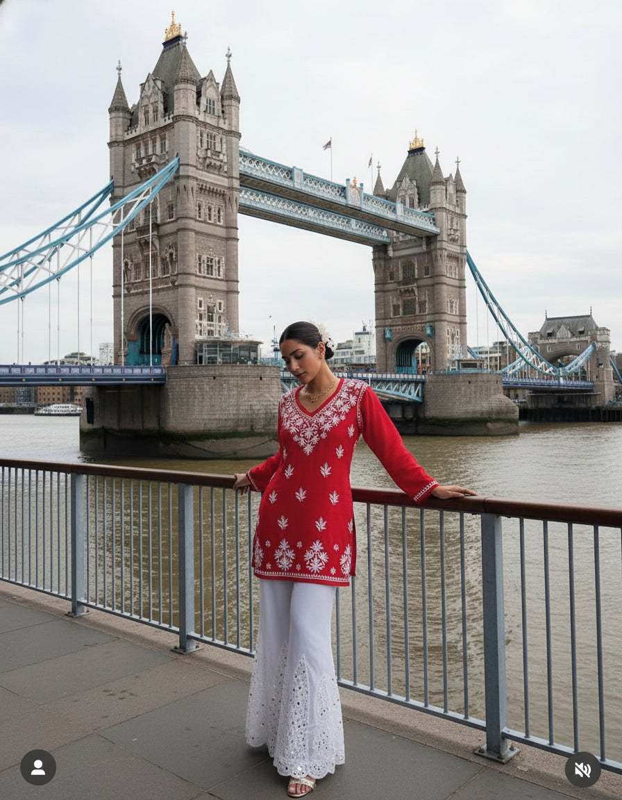 Woman in a red top with white patterns and white pants standing on a bridge with modern buildings in the background.