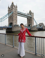 Woman in a red top with white patterns and white pants standing on a bridge with modern buildings in the background.
