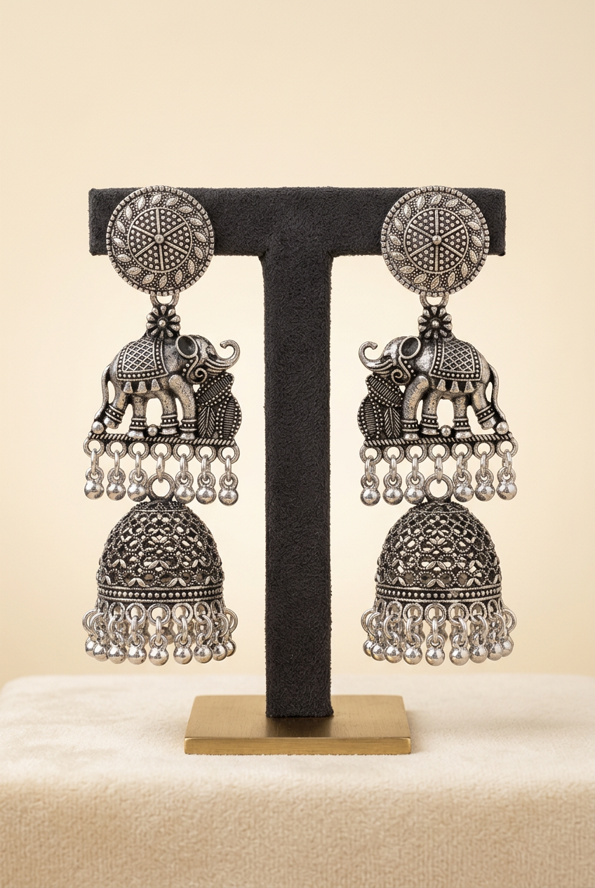 A pair of ornate silver-toned Indian jhumka earrings displayed on a black T-stand against a warm beige background. The earrings feature elephant motifs, beaded fringe, and bell-shaped dangling elements.
