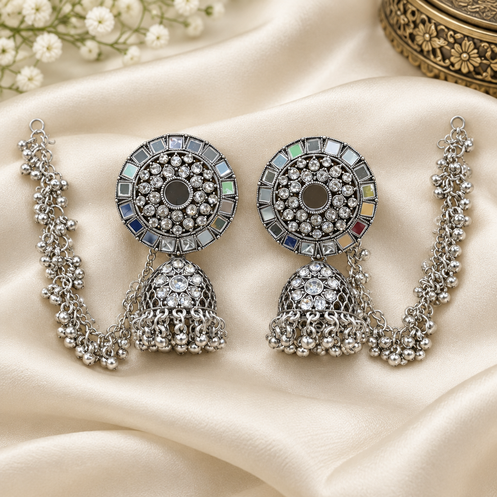 A pair of ornate silver-toned Hare Krishna earrings with multicolored square crystals and dangling chain elements, displayed on draped cream satin fabric.