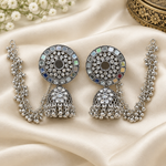 A pair of ornate silver-toned Hare Krishna earrings with multicolored square crystals and dangling chain elements, displayed on draped cream satin fabric.