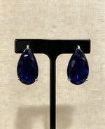 Navy blue teardrop crystal stud earrings in silver-tone three-prong settings on a black stand.