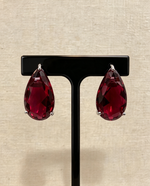 Ruby red teardrop crystal stud earrings in silver-tone three-prong settings on a black stand.