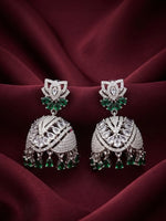 A pair of ornate silver-toned jhumka earrings with green stone accents and dangling teardrop charms, displayed on draped burgundy fabric.