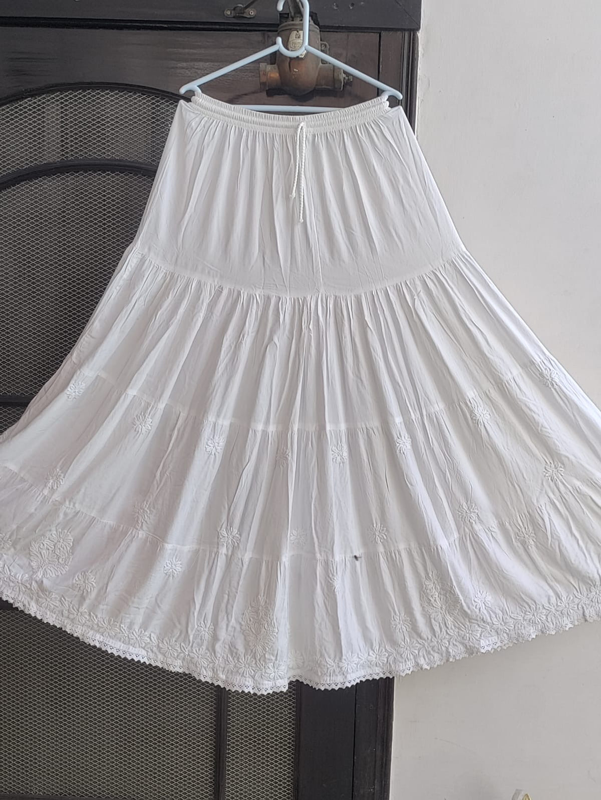 A white tiered skirt with embroidered details and a lace hem, hanging on a hanger against a dark mesh screen and a light wall.